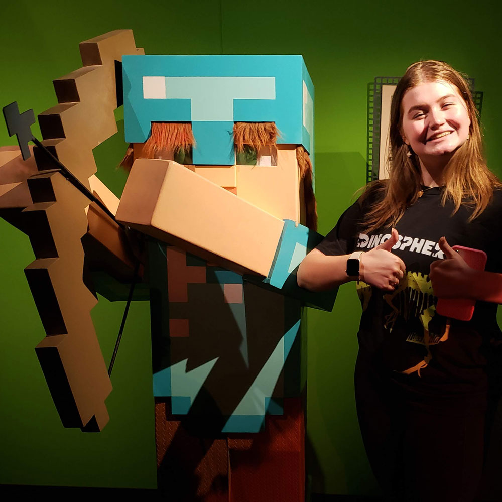 Woman with shoulder-length hair smiling and holding a thumbs up signal while standing next to a life sized sculpture of Alex from Minecraft.